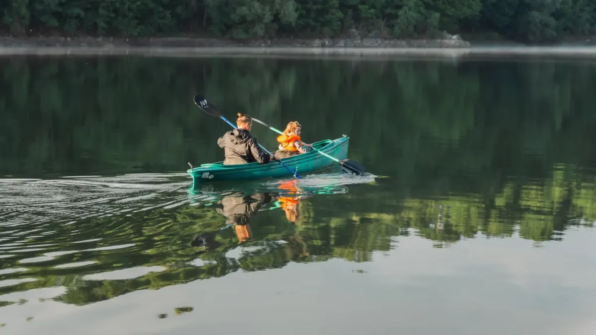 A dog wearing an orange life jacket sitting calmly in the bow of a kayak on calm lake water